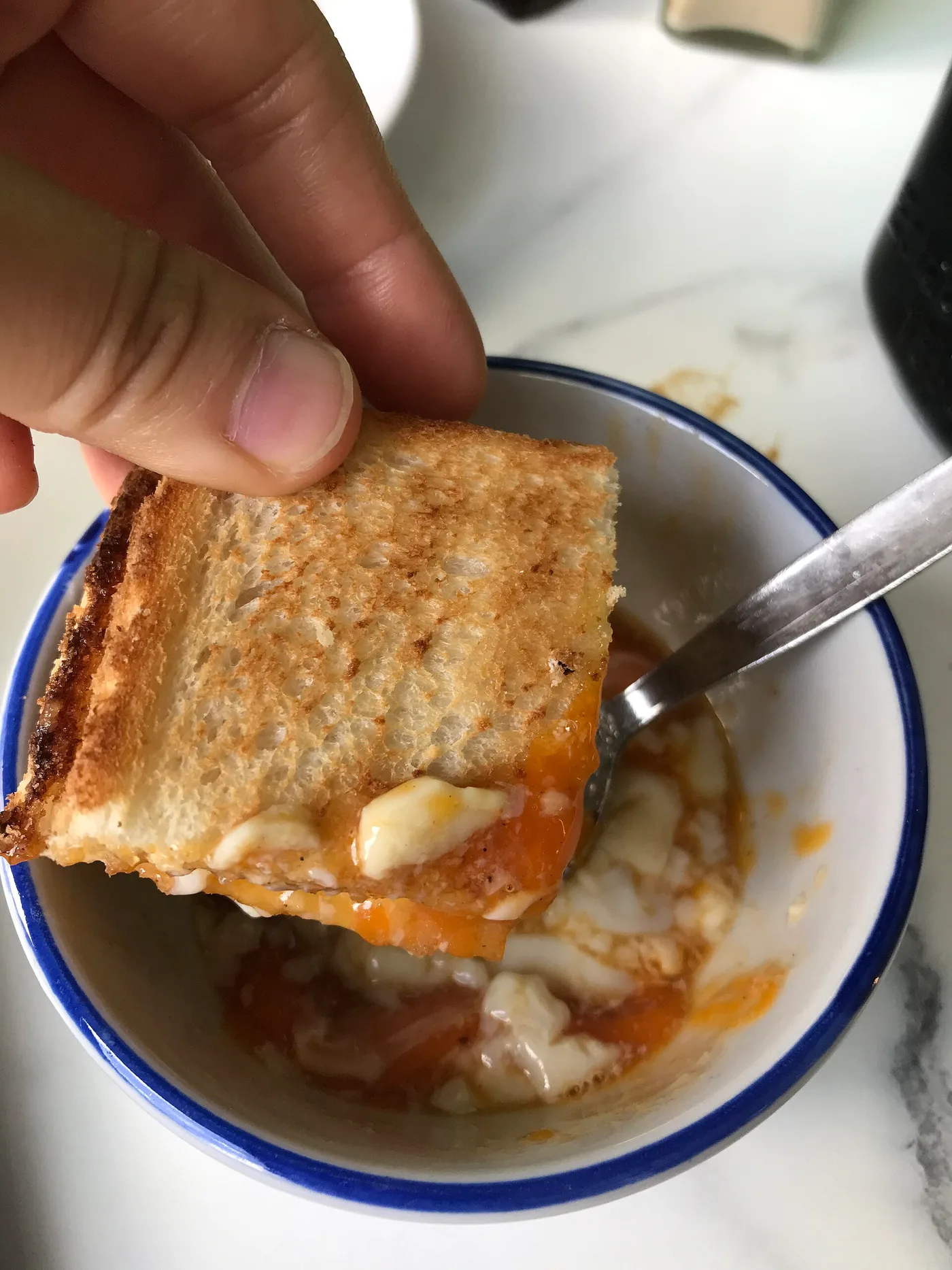 A hand dips a piece of kaya toast into a bowl with a soft-boiled egg in dark soy sauce