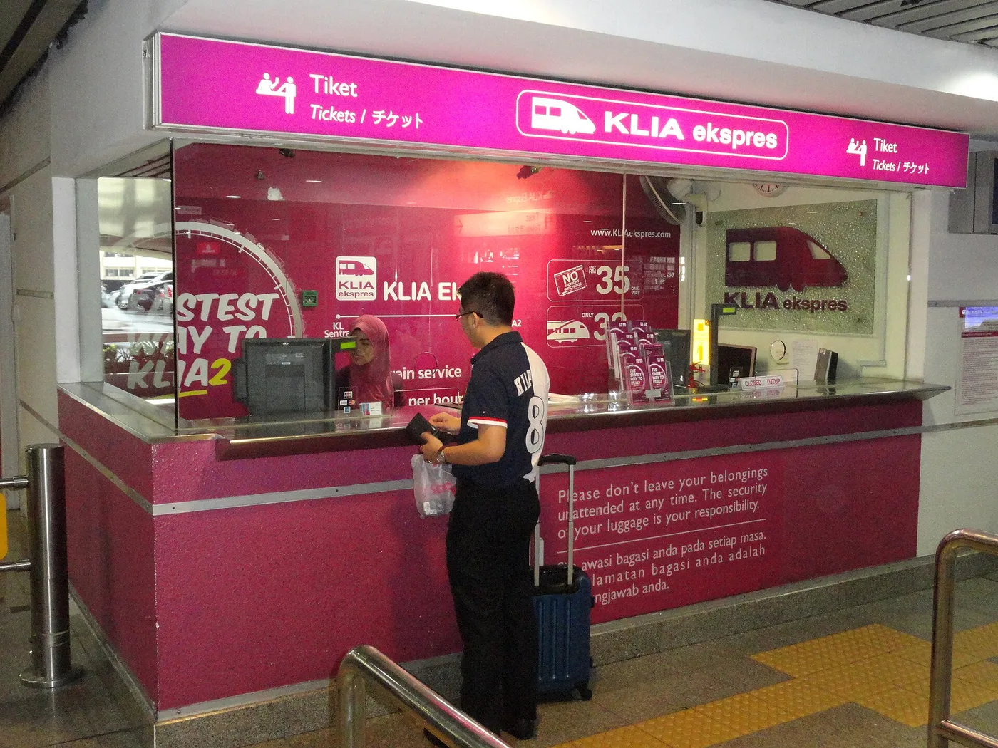 A traveller buying a ticket at the branded KLIA Ekspres counter with a rolling suitcase