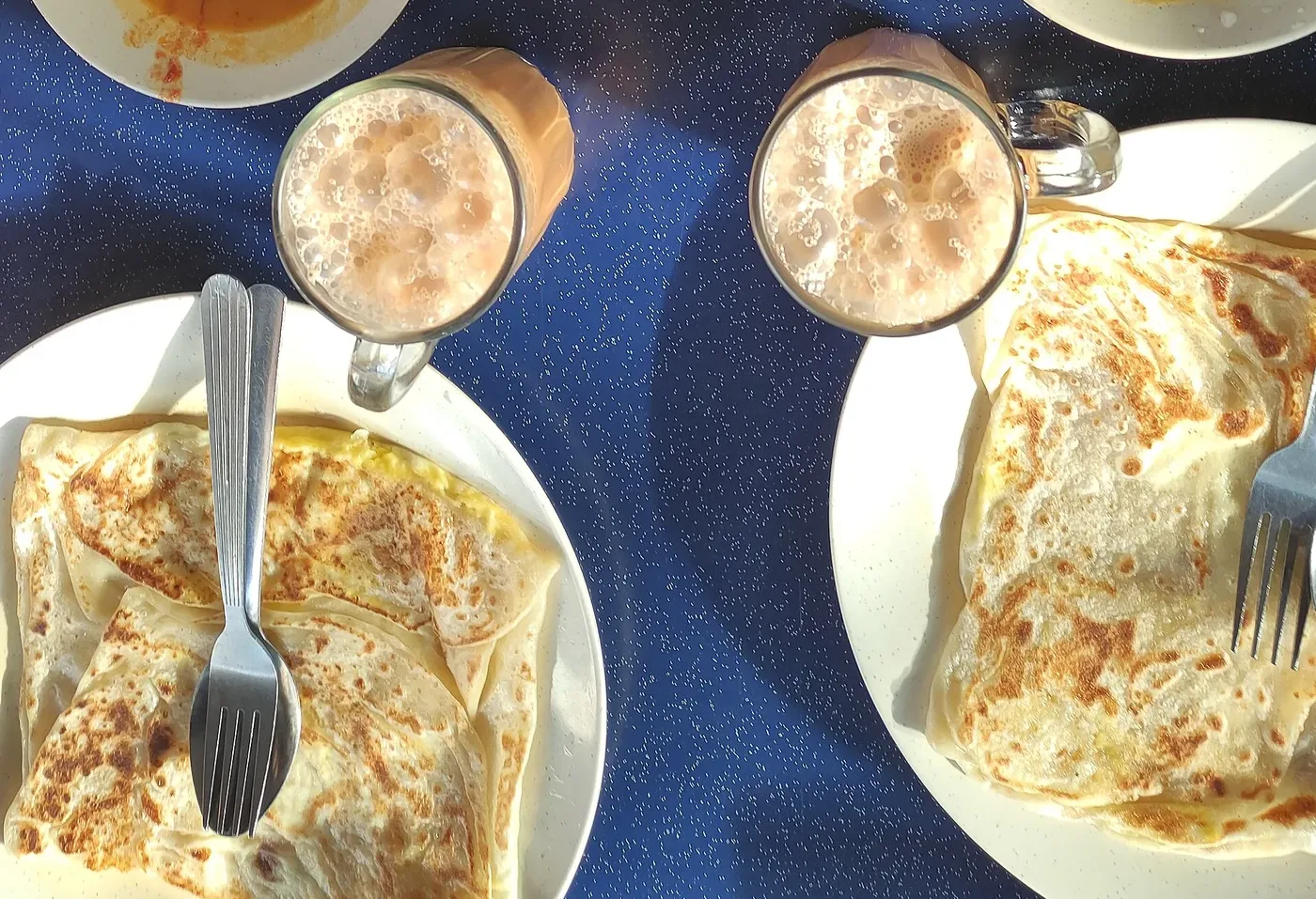 Plates of roti canai with dhal and glasses of frothy teh tarik on a table