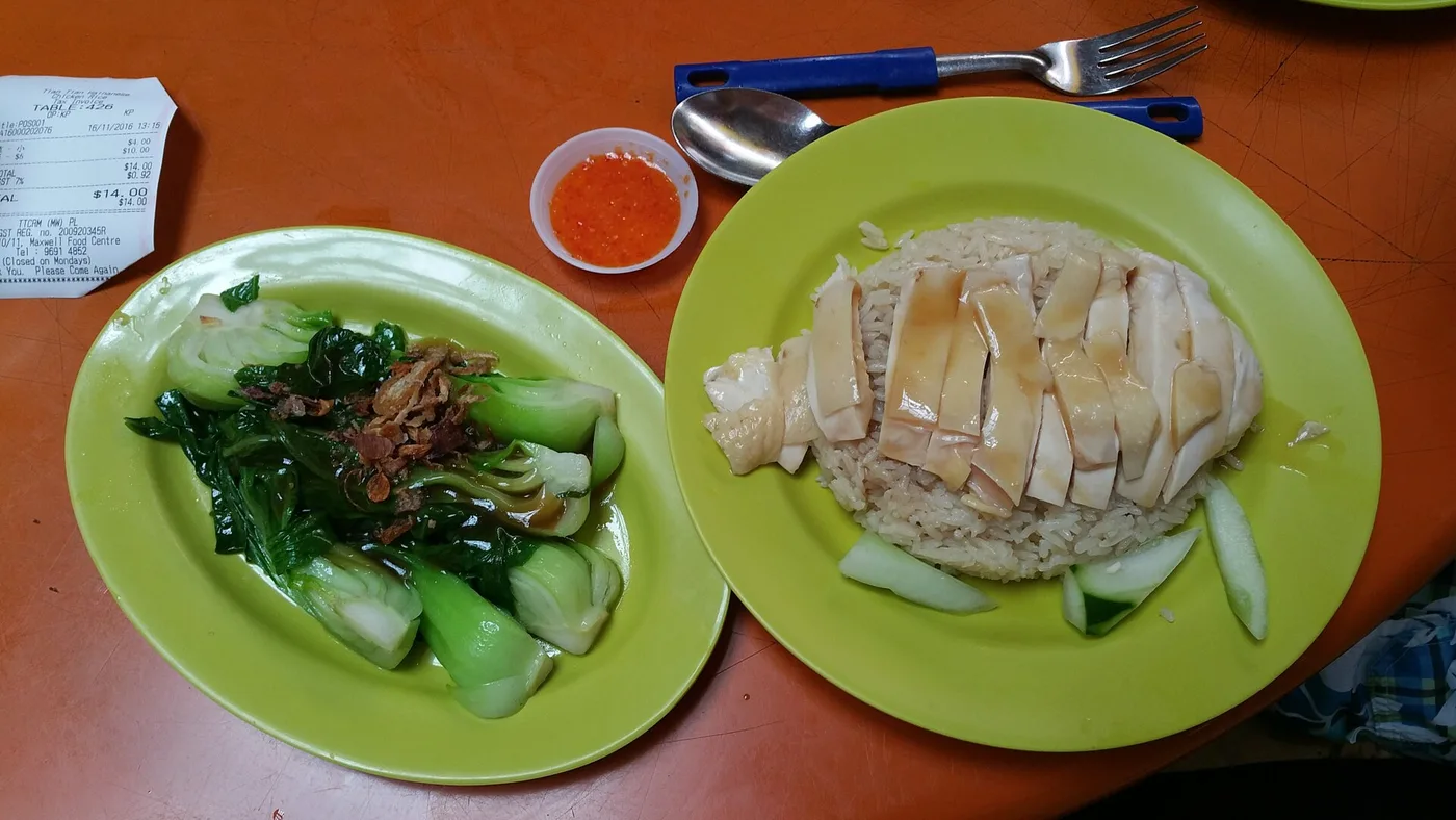 A plate of Hainanese chicken rice with cucumber and greens on an orange hawker table